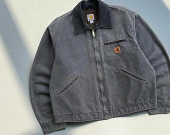 Puede incluir: Chaqueta de trabajo gris con cuello oscuro y cremallera completa. La chaqueta tiene dos bolsillos delanteros y un pequeño parche con el logotipo marrón. La chaqueta está hecha de un material duradero.