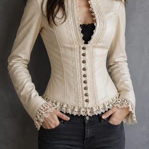 May include: A cream-coloured long-sleeve jacket featuring a high collar, a front button closure, and intricate lace trim along the neckline, cuffs, and hem. The jacket is worn over a black top and paired with dark grey denim jeans.
