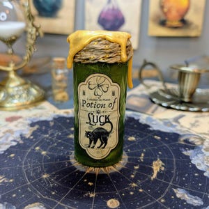 Op de afbeelding: Een cilindervormige groene glazen fles met een etiket waarop Potion of LUCK staat, voorzien van een klavertjevier en een zwarte kat. De bovenkant is verzegeld met natuurlijk touw en druipende gele was voor een textuurrijk uiterlijk.