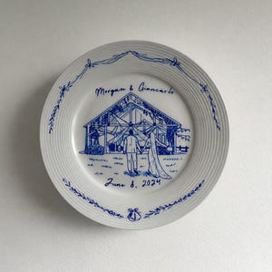 Puede incluir: Plato de cerámica blanco con una ilustración azul de un granero y una pareja. El plato está decorado con detalles florales azules y los nombres "Morgan & Giancarlo" y la fecha "8 de junio de 2024".