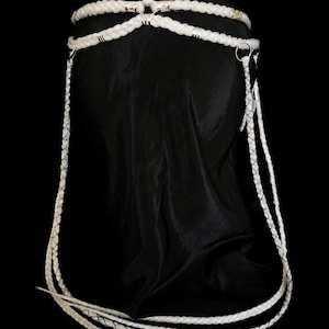 May include: A white braided cord headpiece featuring multiple interconnected strands and silver-toned metal accents. The design includes a circular crown structure with long, dangling braided cords that extend downwards, secured by small metallic rings.