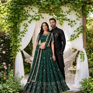 May include: A couple poses in a garden setting, showcasing formal attire. The woman wears a dark green, embellished lehenga with a matching dupatta. The man is in a black, embroidered suit. The background includes greenery and white draping.