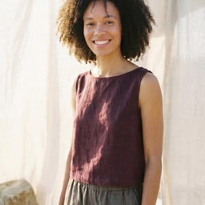 May include: A sleeveless, burgundy-coloured top with a boat neck. The top is paired with grey trousers. The person in the image is smiling. The background is a light-coloured fabric.