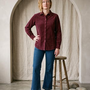 May include: A long-sleeved, button-down shirt in a deep burgundy corduroy fabric with a classic collar, paired with dark blue denim jeans that feature a straight-leg cut and hemmed bottoms, worn with dark brown leather oxford shoes.