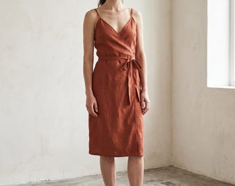 May include: A rust-colored wrap dress with spaghetti straps and a matching waist tie. The knee-length dress is made of a lightweight fabric. The model is wearing brown leather clogs. The dress is suitable for casual or semi-formal occasions.