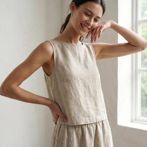 Puede incluir: Un top sin mangas de lino beige y pantalones a juego. El top tiene un cuello redondo simple. Los pantalones tienen cintura fruncida. El conjunto está hecho de una tela ligera y texturizada. La modelo sonríe.