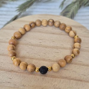 Könnte beinhalten: Ein elastisches Armband aus glatten, hellbraunen Holzperlen, die mit kleinen, polierten goldfarbenen Metallkugeln durchsetzt sind. Eine einzelne, texturierte schwarze Lavasteinperle befindet sich in der Mitte und bildet den Blickfang.