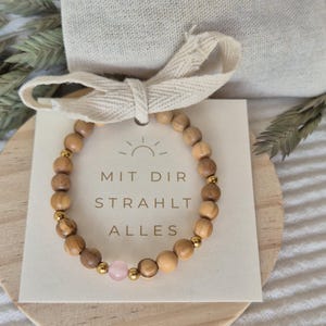 Könnte beinhalten: Ein Perlenarmband aus runden Holzperlen, kleinen goldfarbenen Metallzwischenelementen und einer einzelnen hellrosa Perle. Das Armband ist auf einer quadratischen weißen Karte mit dem Text MIT DIR STRAHLT ALLES und einem Sonnensymbol befestigt.