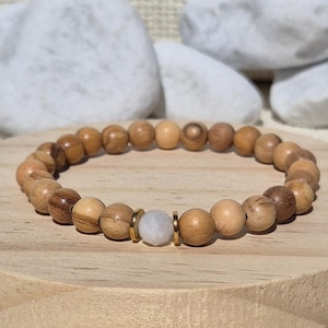 Könnte beinhalten: Ein Perlenarmband aus glatten, runden, hellbraunen Holzperlen, die in einem Kreis angeordnet sind. Das Armband enthält eine einzelne, mittig platzierte, facettierte, durchscheinende weiße Steinperle, die von zwei dünnen, flachen, goldfarbenen Metallabstandshaltern flankiert wird.