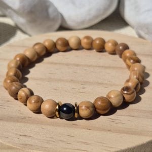 Könnte beinhalten: Ein elastisches Armband aus runden, hellbraunen Holzperlen mit sichtbarer Maserung. Das Design zeichnet sich durch eine einzelne, glatte, glänzend schwarze Perle in der Mitte aus, die von zwei dünnen, flachen, goldfarbenen Metallzwischenscheiben flankiert wird.