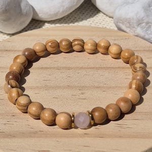 Könnte beinhalten: Ein Perlenarmband mit runden, hellbraunen Holzperlen mit sichtbarer Maserung. Das Armband enthält eine einzelne, facettierte, blassrosa Edelsteinperle, die zwischen zwei kleinen, flachen, goldfarbenen Metall-Abstandshaltern zentriert ist.