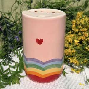 Könnte beinhalten: Ein rosa Keramikbecher mit einem roten Herzen und einem Regenbogen-Chevron-Design. Die Oberseite hat kleine rote Herzakzente. Der Becher ist zylindrisch und scheint handgefertigt zu sein.
