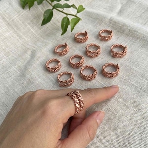 May include: Copper-colored rings with a braided design are displayed on a neutral linen surface. One ring is worn on a finger, showcasing the intricate weave. Several other rings are scattered around, highlighting the jewelry's craftsmanship.