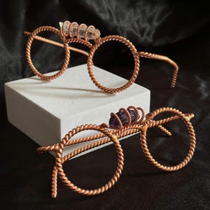 Könnte beinhalten: Zwei Paar handgefertigte Brillen aus Kupferdraht mit runden Fassungen. Ein Paar hat einen klaren Kristallakzent, das andere einen violetten Amethysten. Die Fassungen haben ein gedrehtes Seildesign.