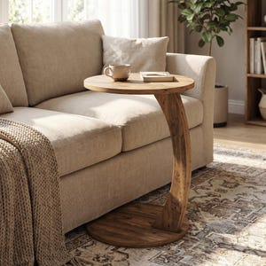 May include: A wooden side table with a round top and a curved leg. The table is made of light brown wood and is placed next to a beige sofa. A cup and a book are on the table.