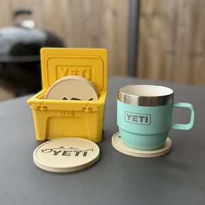 Könnte beinhalten: Ein leuchtend gelber Miniatur-Kühlbehälter mit runden beigen Untersetzern, die das YETI-Logo und eine Fischgrafik zeigen. Daneben steht ein hell mintgrüner Isolierbecher mit silbernem Rand und YETI-Logo auf einem passenden beigen Untersetzer.