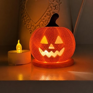 May include: A bright orange jack-o'-lantern pumpkin with a textured, knit-like surface and a dark stem. The pumpkin features triangular eyes and a jagged mouth, illuminated from within by a warm light, sitting next to a small white tea light.