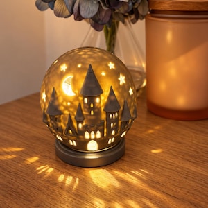 May include: A spherical ceramic lamp featuring a cutout design of a castle with towers, a crescent moon, and stars. The lamp emits a warm, glowing light through the openings, casting patterns onto the surface below, and sits on a circular base.