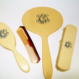 May include: A set of four ivory-colored grooming tools with black monogrammed initials. The set includes a hand mirror, a hairbrush, a comb, and a clothes brush.