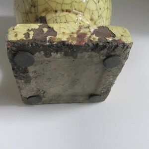 May include: A yellow ceramic vase with a crackled glaze and four black rubber feet on the bottom.