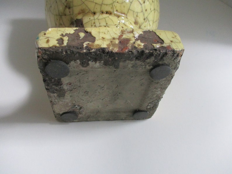 May include: A yellow ceramic vase with a crackled glaze and four black rubber feet on the bottom.