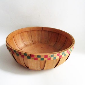 May include: A round, light-colored wooden basket with a woven rim featuring a pattern of red and green squares. The basket is empty and sits on a white surface, showcasing its natural wood grain.