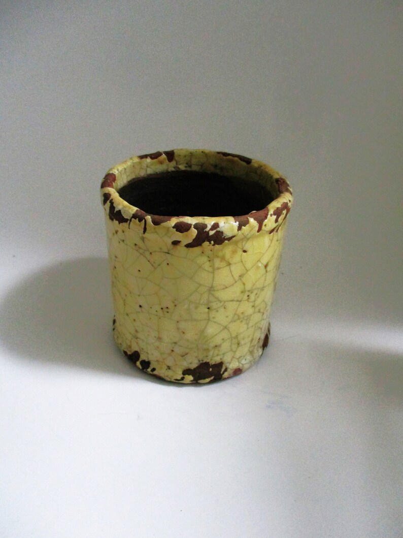 May include: A yellow ceramic planter with a crackled glaze and brown accents. The planter has a round opening and a cylindrical shape.