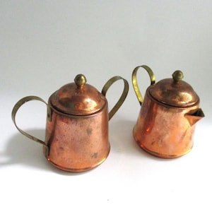 May include: Two copper vessels with brass handles and lids. One vessel features a small spout, while the other has two handles on opposite sides. Both items have a metallic finish with visible signs of wear and oxidation on the copper surfaces.