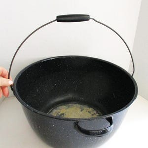 May include: A black speckled enamelware pot with a wire handle and side handles. The pot has a rounded shape and a black interior. The handle is black with a black grip. The pot is likely used for cooking or storage.