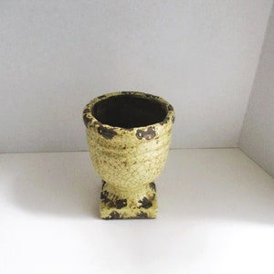 May include: A small, yellow ceramic planter with a brown glaze and a square base. The planter has a crackled finish and a distressed look.