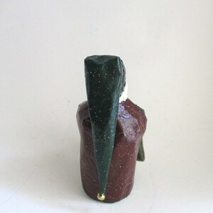 May include: A green and red paper mache figurine of a person wearing a green hat with a gold accent. The figurine is standing on a white surface.