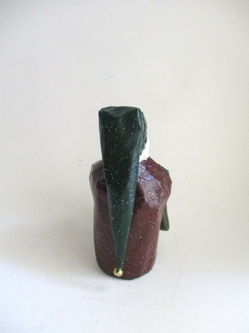 May include: A green and red paper mache figurine of a person wearing a green hat with a gold accent. The figurine is standing on a white surface.