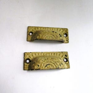 May include: Two rectangular metal drawer pulls with a distressed pale yellow finish. Each pull features a curved handle section with a raised, embossed semi-circular pattern and two circular screw holes on the flat mounting plates at each end.