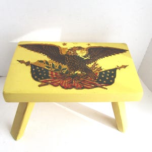 May include: Yellow wooden step stool featuring a patriotic design. The top displays a detailed image of an eagle with outstretched wings, the American flag, and stars. The stool has three legs.