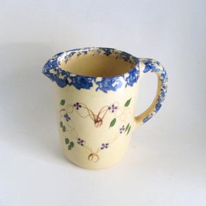 May include: Cream-colored ceramic pitcher with a blue floral rim and handle. The pitcher features a heart-shaped vine design with blue flowers and green leaves. The pitcher has a spout for easy pouring.