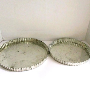 May include: Two round, silver-colored tart pans with fluted edges. The pans are made of metal and have a slightly worn appearance. One pan is larger than the other. The pans are empty and ready for baking.