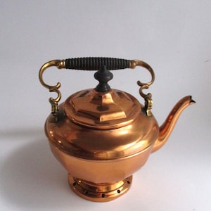 May include: A copper teapot with a black handle and brass accents. The teapot has a round body and a lid with a small knob on top. The teapot is sitting on a white surface.
