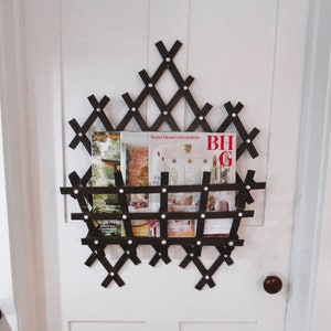 May include: A dark brown wooden magazine rack with a geometric design, mounted on a white door. The rack holds several magazines, including one with "Better Homes & Gardens" on the cover. White dots secure the wooden pieces.