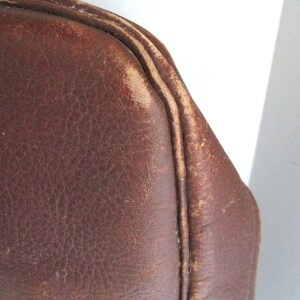 May include: Close-up of a brown leather bag corner. The leather has a distressed appearance with visible wear and tear, showcasing a vintage aesthetic. The stitching is visible along the edges, adding to the bag's character. The bag's color is a rich, warm brown.
