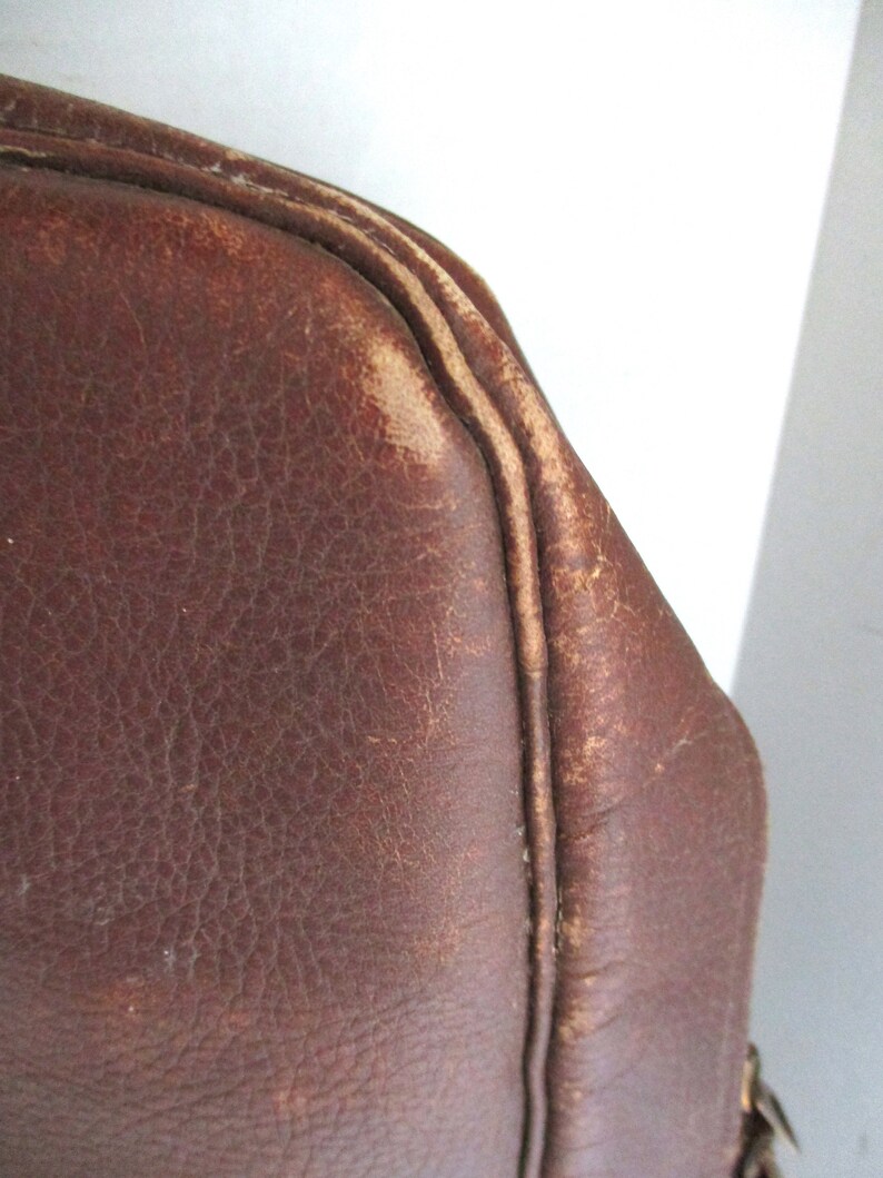 May include: Close-up of a brown leather bag corner. The leather has a distressed appearance with visible wear and tear, showcasing a vintage aesthetic. The stitching is visible along the edges, adding to the bag's character. The bag's color is a rich, warm brown.