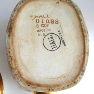 May include: Base of a vintage Hall ceramic teapot, off-white with brown edging. The text "HALL 01088 6 CUP MADE IN U.S.A." is printed in brown. The handle is gold-coloured.