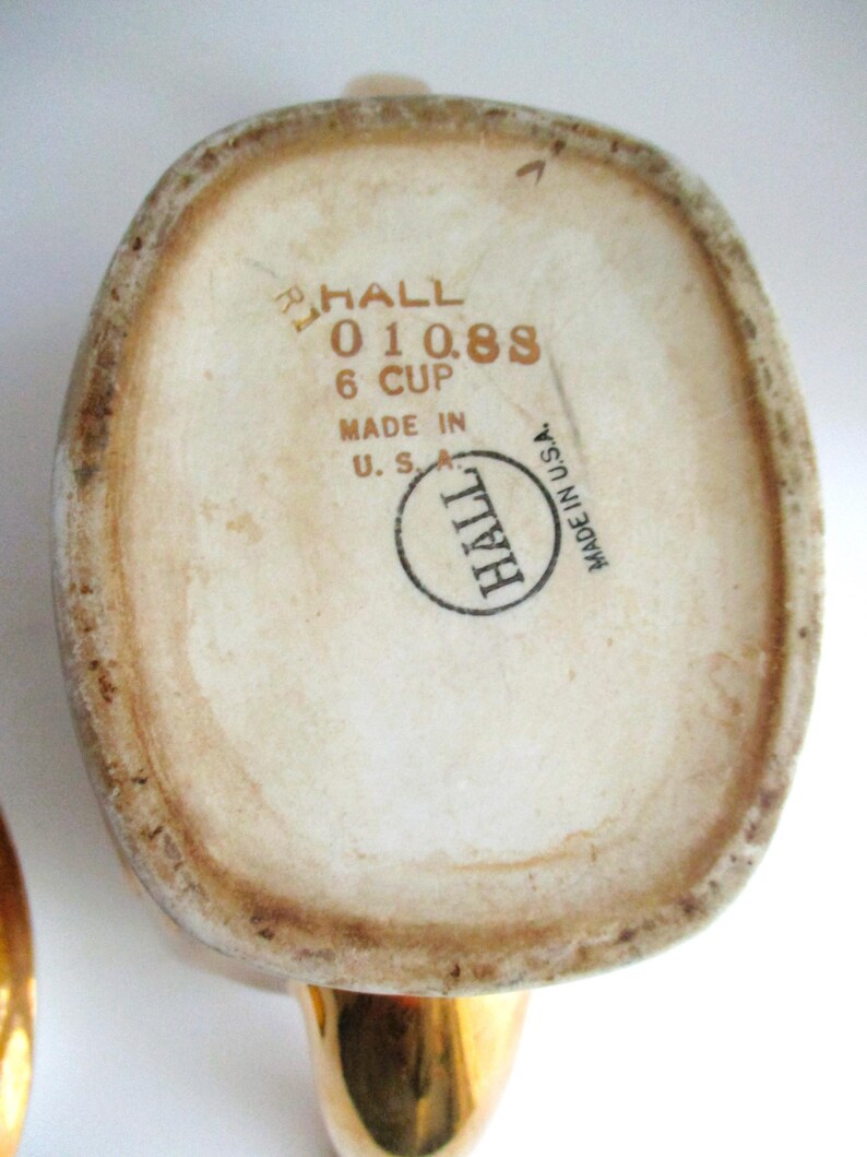 May include: Base of a vintage Hall ceramic teapot, off-white with brown edging. The text "HALL 01088 6 CUP MADE IN U.S.A." is printed in brown. The handle is gold-coloured.