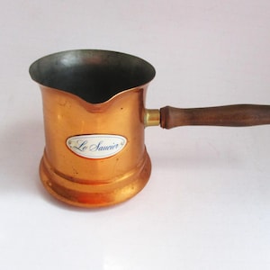 May include: A copper-colored coffee pot with a wooden handle. The pot has a rounded base and a flared rim. The label on the pot reads "Le Saucier". The handle is attached with a brass fitting. The pot is designed for stovetop use.