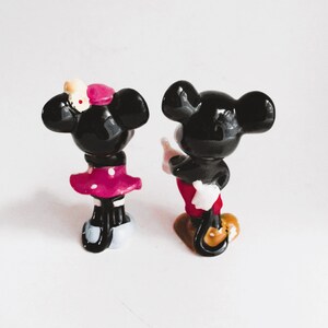 May include: A black and white figurine of Mickey Mouse and Minnie Mouse. Mickey is wearing red shorts and white gloves. Minnie is wearing a pink polka dot dress and a yellow bow.