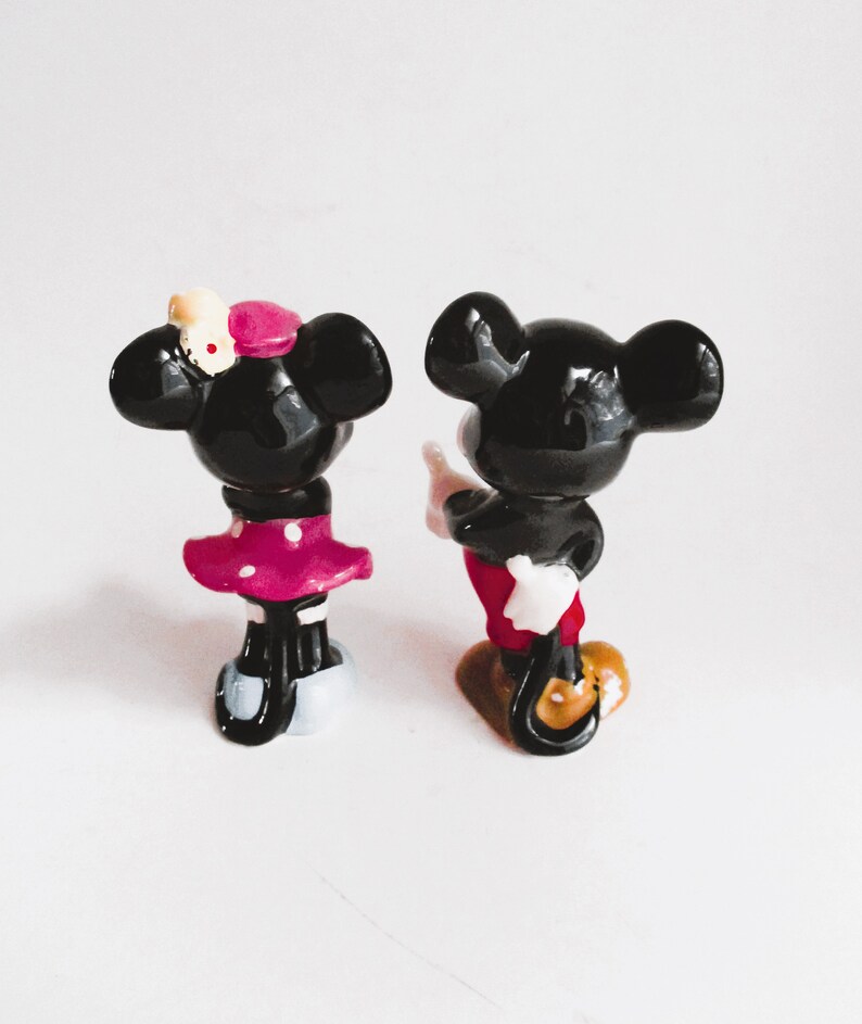 May include: A black and white figurine of Mickey Mouse and Minnie Mouse. Mickey is wearing red shorts and white gloves. Minnie is wearing a pink polka dot dress and a yellow bow.