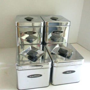 May include: A set of four stainless steel kitchen canisters with black accents. Each canister is labeled with the words "flour", "sugar", "coffee", and "tea" in a black oval. The lids have a black square handle.