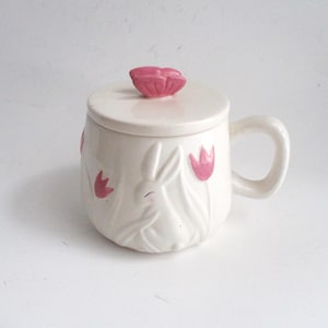 May include: A white ceramic mug with a lid, featuring a bunny and tulip design. The mug has a handle and is decorated with pink tulips. The lid is topped with a pink flower. A charming Easter-themed mug.