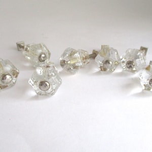 May include: A collection of eight clear glass cabinet knobs with a faceted, hexagonal design. Each knob features a silver-colored metal base and a central, rounded silver accent. The knobs are arranged on a white surface.