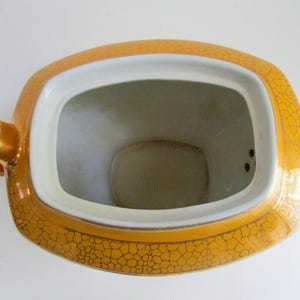 May include: Overhead shot of a vintage teapot. The teapot is a golden yellow colour with a cracked pattern design. The interior is white. The teapot has a rectangular opening and a gold handle.