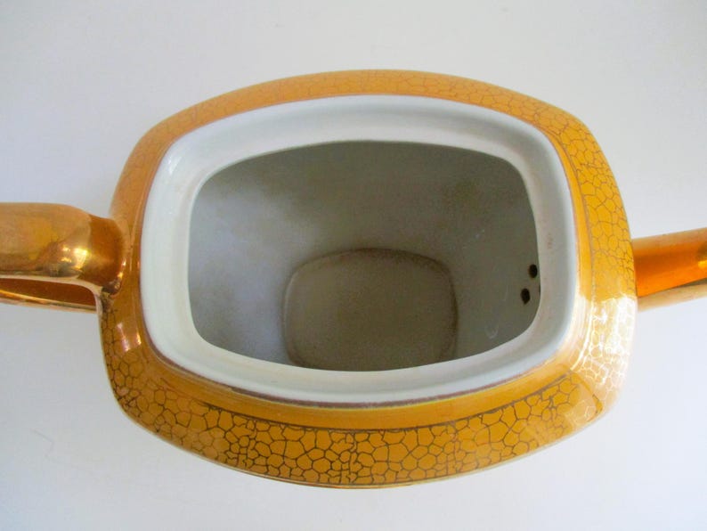 May include: Overhead shot of a vintage teapot. The teapot is a golden yellow colour with a cracked pattern design. The interior is white. The teapot has a rectangular opening and a gold handle.